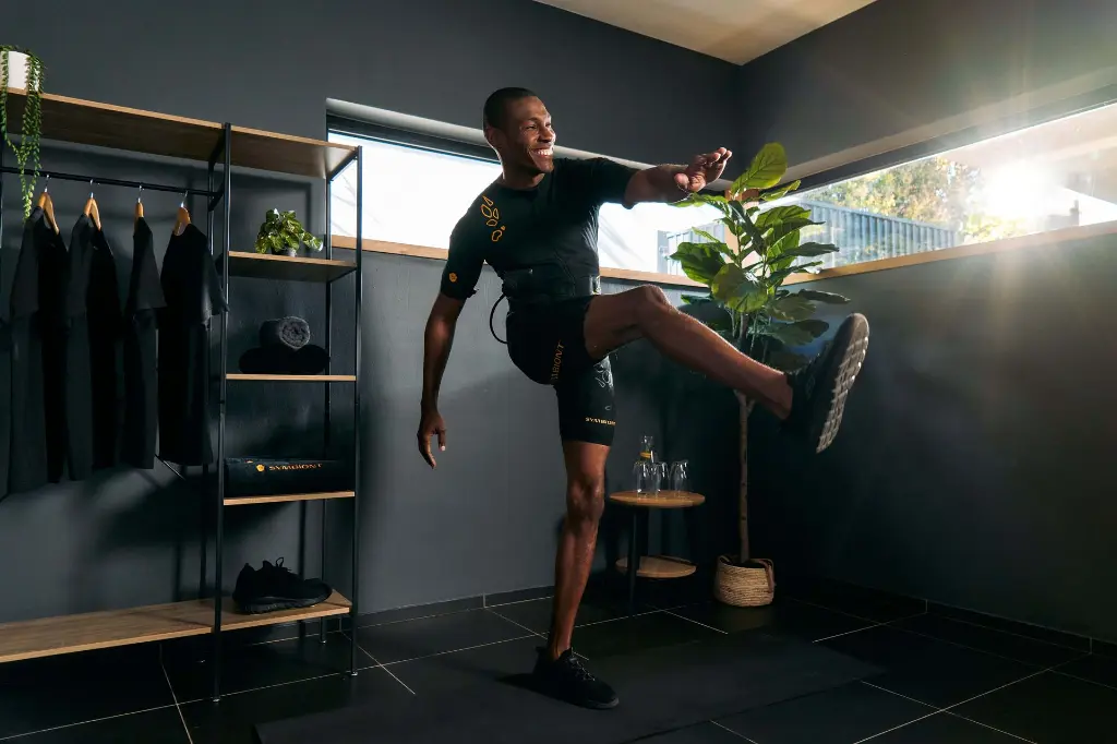 SYMBIONT EMS Experience – The Future of Fitness with Advanced Electrical Muscle Stimulation A man in the SYMBIONT EMS suit is performing a leg lift exercise. The room is filled with natural light. The image represents the future of EMS training and fitness.