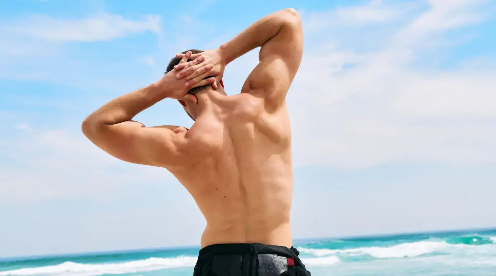 A muscular man seen from behind as he stretches with his hands behind his head. He stands on a beach with waves and a blue sky in the background. The image highlights his strong, well-defined back.